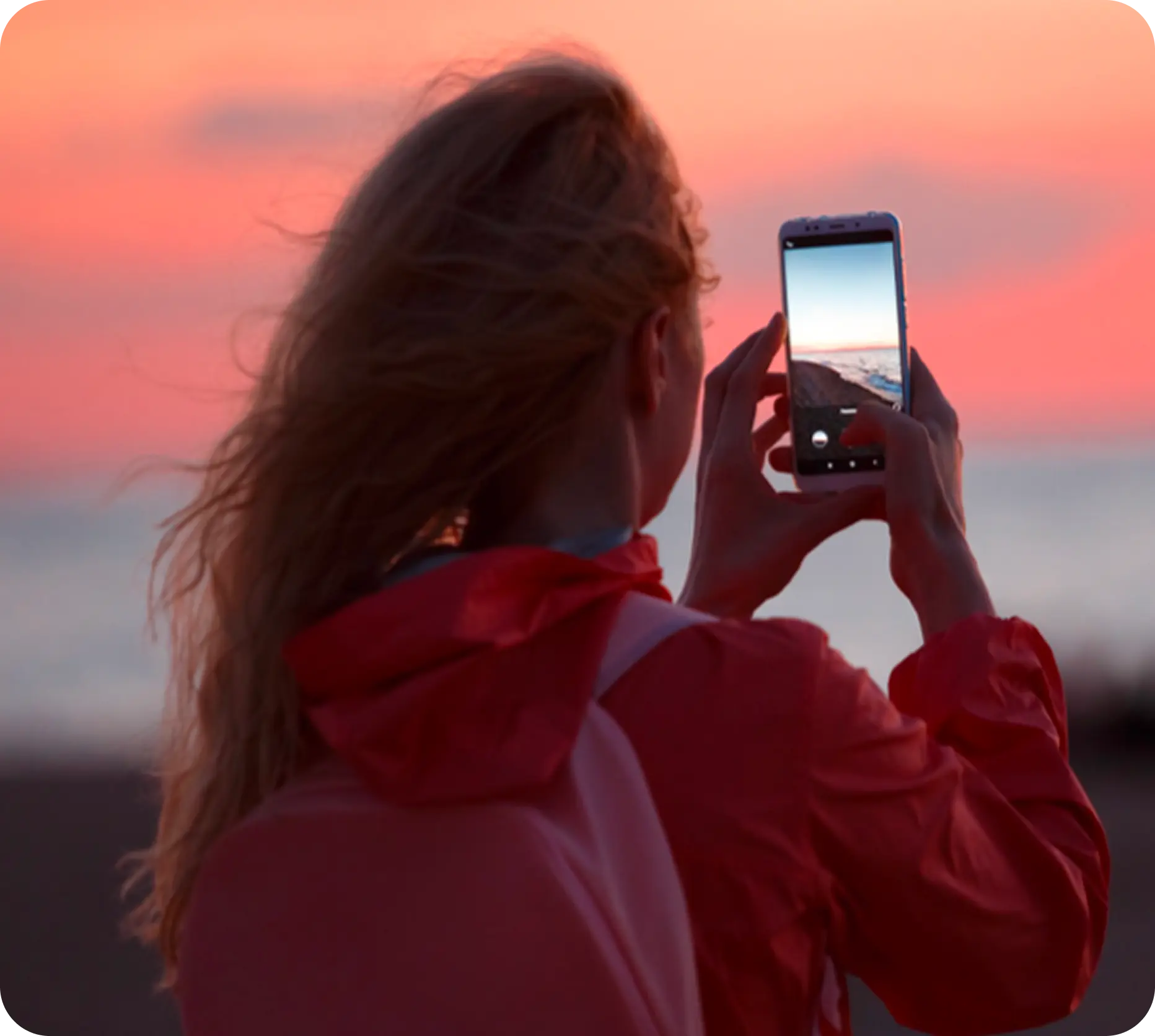 Woman in a red jacket taking a photo of the ocean at sunset with her smartphone